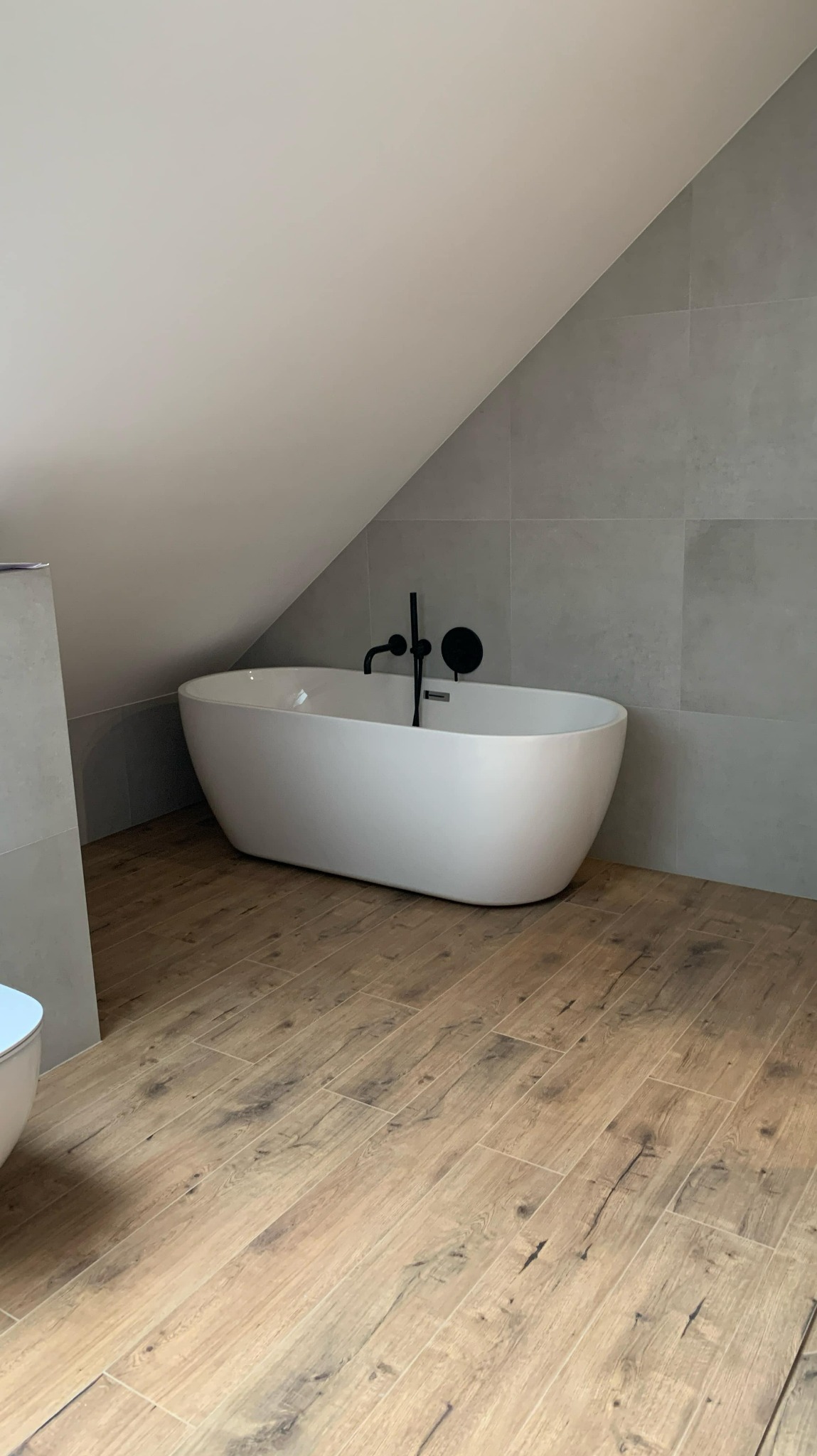 Nowoczesna biała wanna wolnostojąca z czarną armaturą w łazience na poddaszu z drewnopodobną podłogą i szarymi płytkami ściennymi.