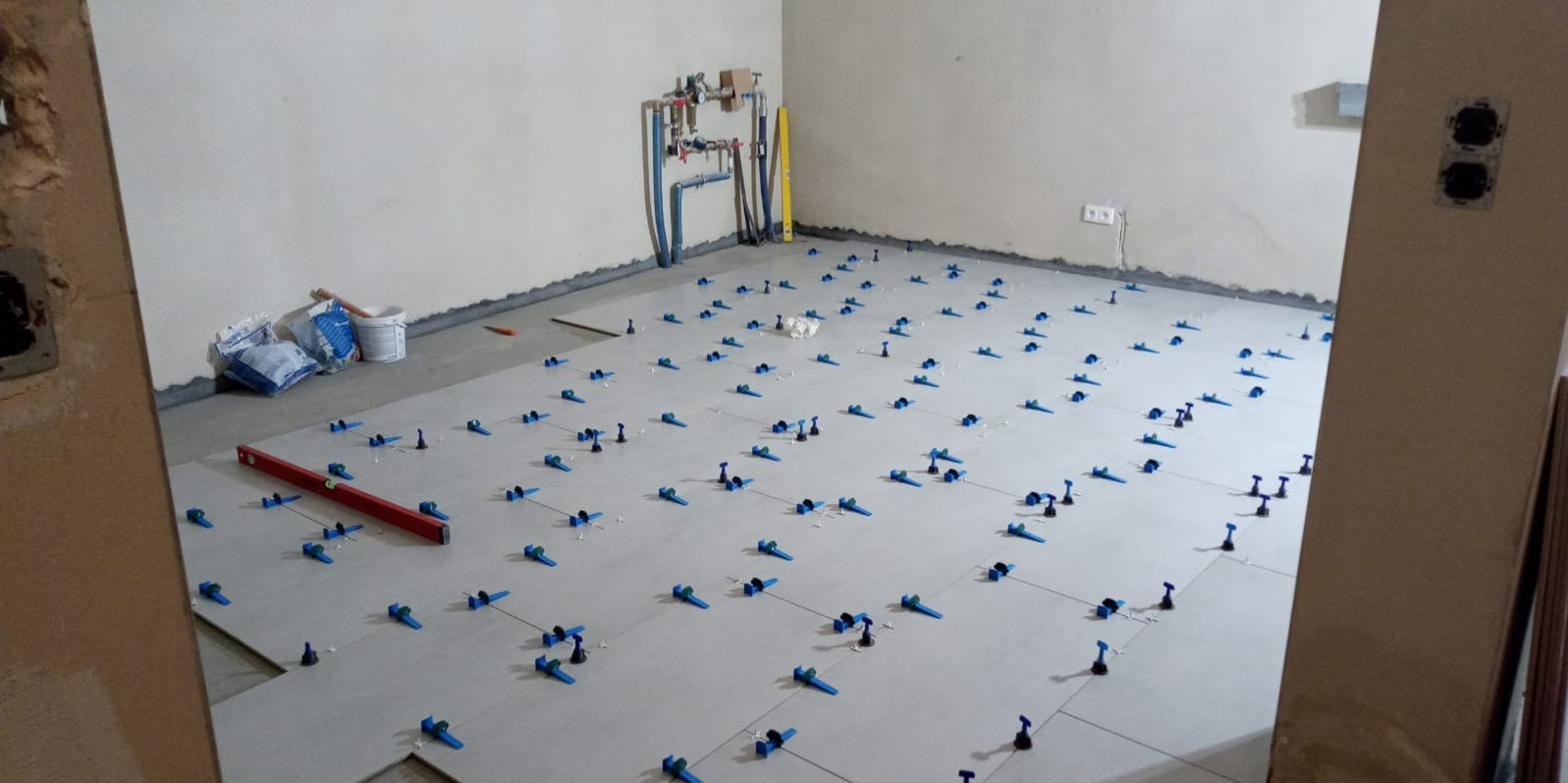 Układanie jasnych płytek podłogowych z użyciem niebieskich klipsów poziomujących w trakcie prac wykończeniowych; widoczne worki z zaprawą i poziomica.