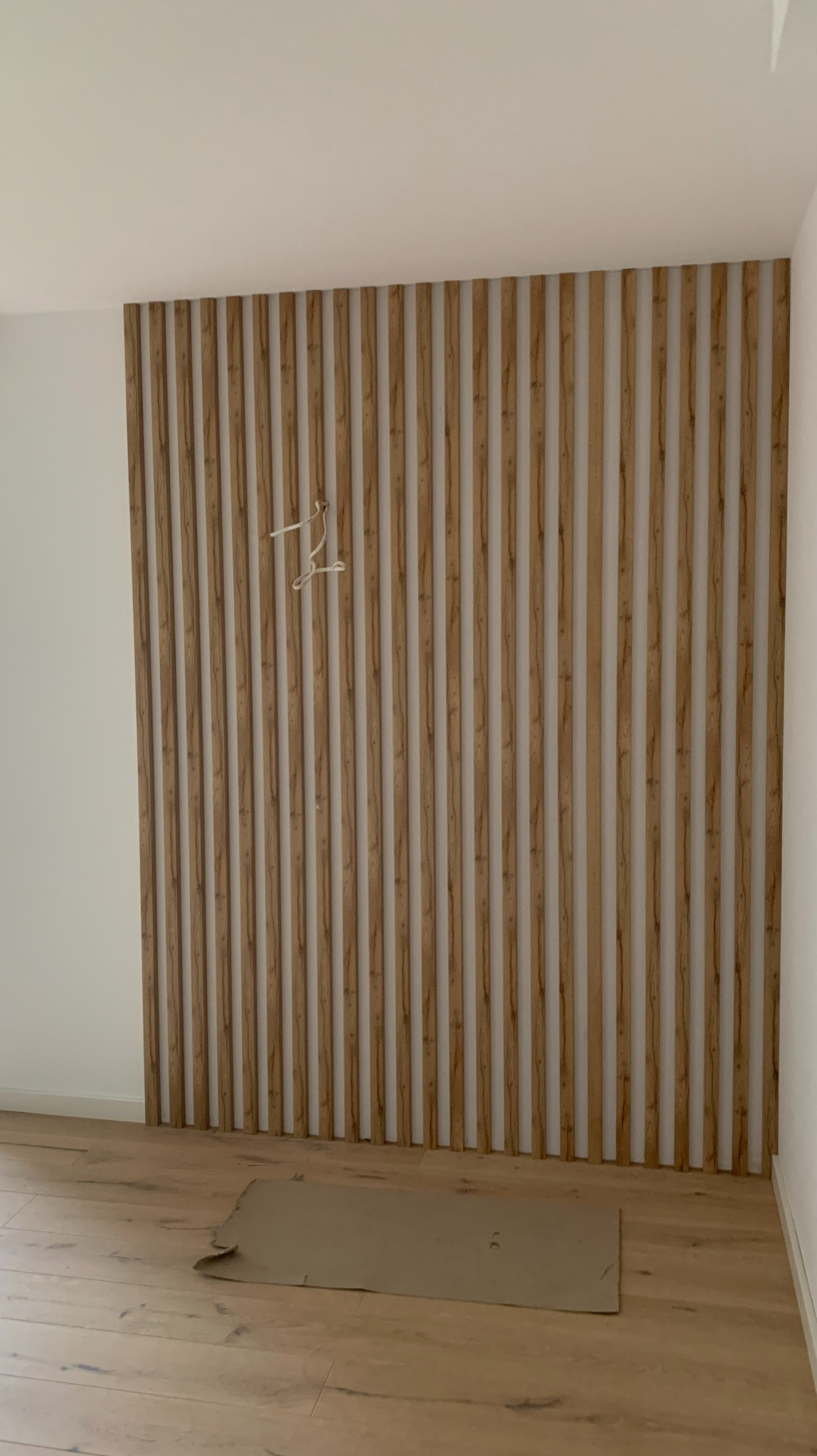 Drewniane lamele dekoracyjne na ścianie w trakcie montażu, widoczna podłoga i karton ochronny, na lamelach wisi biały wieszak.