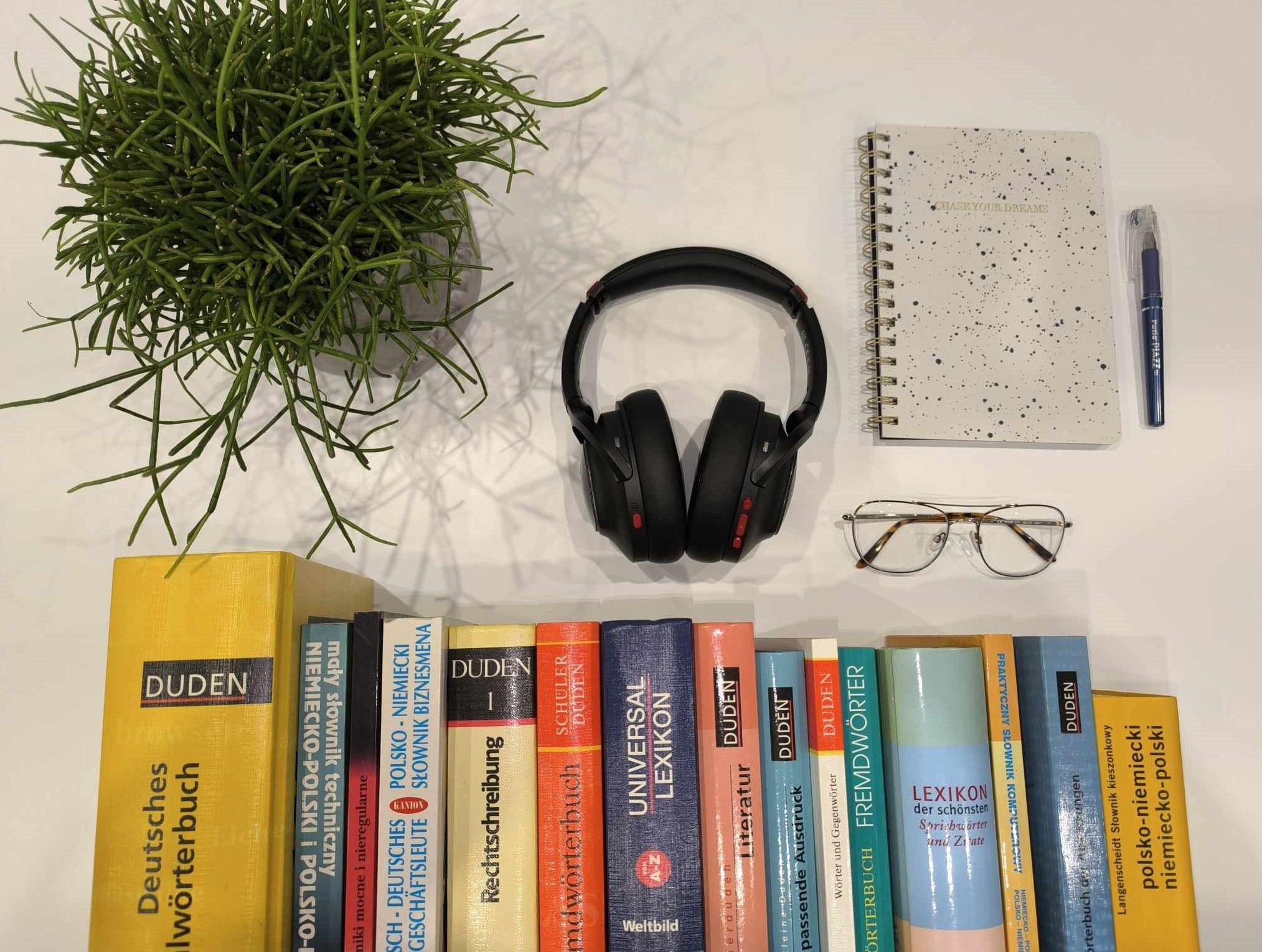 Rząd słowników niemieckich i polsko-niemieckich, notatnik z napisem 'Chase your dreams', słuchawki, okulary i zielona roślina na białym tle.