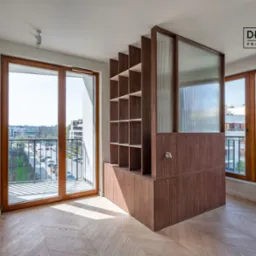 Wnętrze mieszkania z drewnianą zabudową meblową oddzielającą kuchnię od salonu, z otwartymi półkami i przeszkleniem, widok na miasto z balkonu.