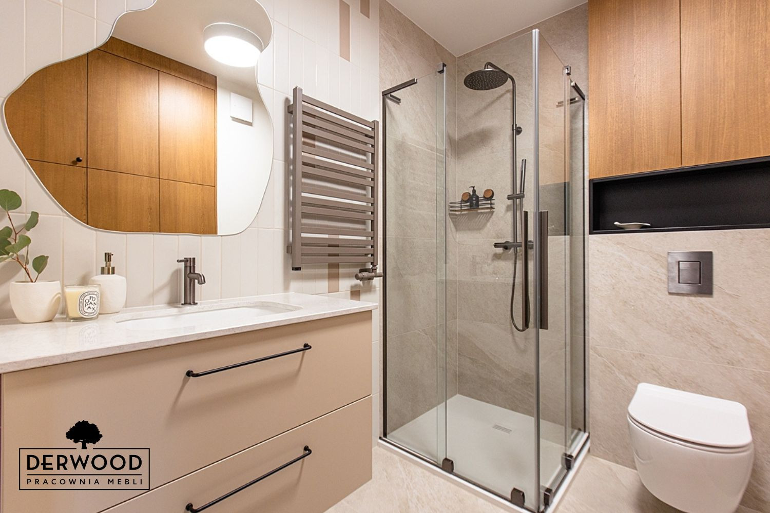 Nowoczesna łazienka z beżową szafką pod umywalką z czarnymi uchwytami, asymetrycznym lustrem i kabiną prysznicową ze szkła, widoczne logo producenta mebli.