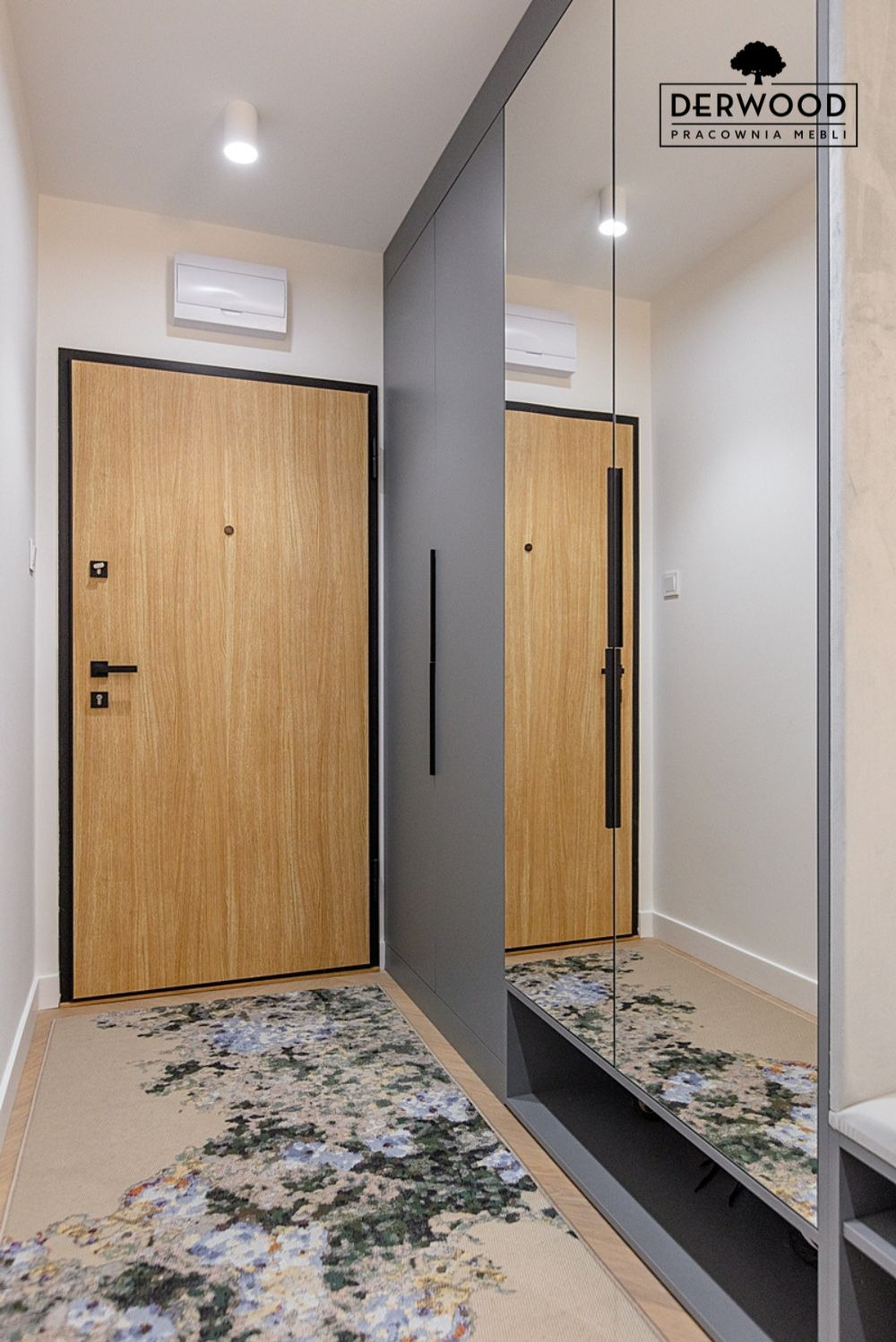 Wnętrze przedpokoju z drewnianymi drzwiami z czarną ramą, szarą szafą z lustrem i wzorzystym dywanem, widoczne logo firmy Derwood Pracownia Mebli w odbiciu lustrzanym.