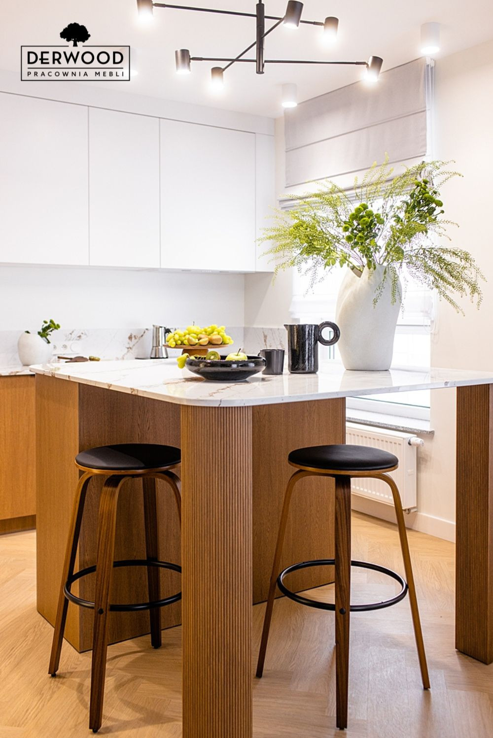 Nowoczesna wyspa kuchenna z marmurowym blatem, drewnianymi kolumnami z frezowaniem i dwoma drewnianymi hokerami z czarnymi siedziskami, uzupełniona owocami i wazonem z zielonymi gałązkami.