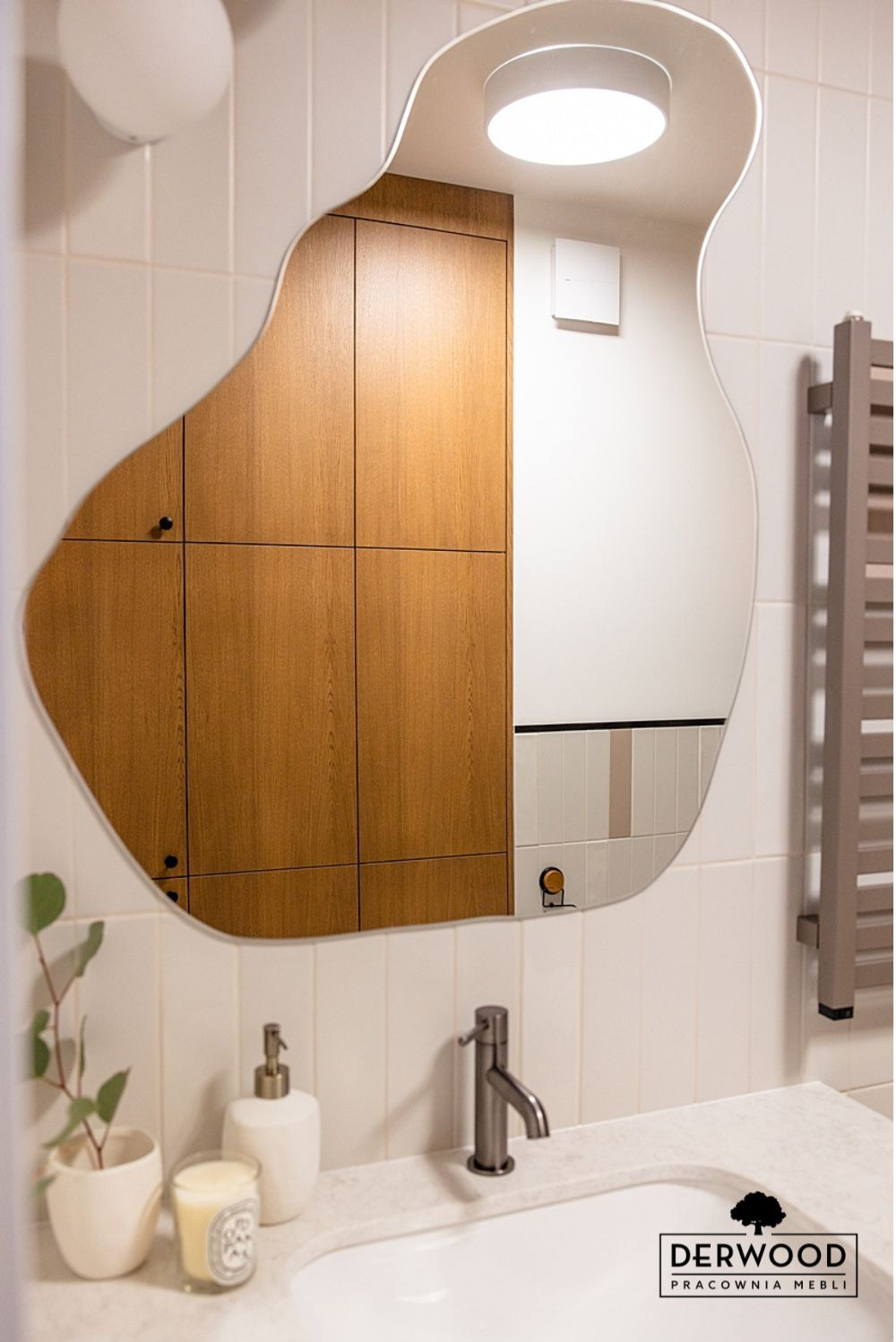 Łazienka z szafką w zabudowie odbijającą się w asymetrycznym lustrze, widoczna umywalka z baterią, dozownik do mydła i roślina w doniczce.