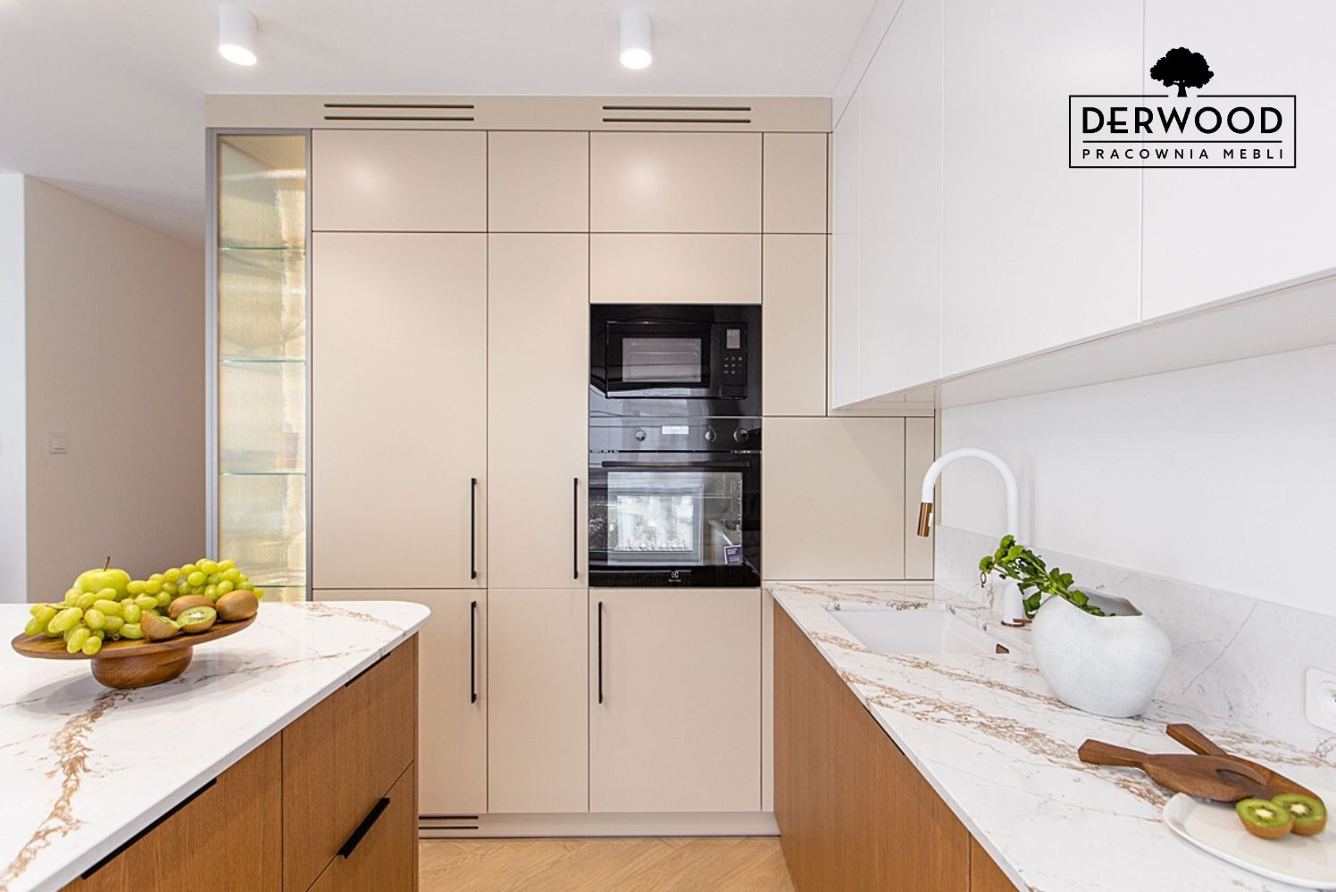 Nowoczesna zabudowa kuchenna w Olecku z beżowymi frontami i czarnymi uchwytami, uzupełniona blatem z marmurowym wzorem i drewnianymi akcentami, oświetlona punktowo, na blacie misa z owocami.