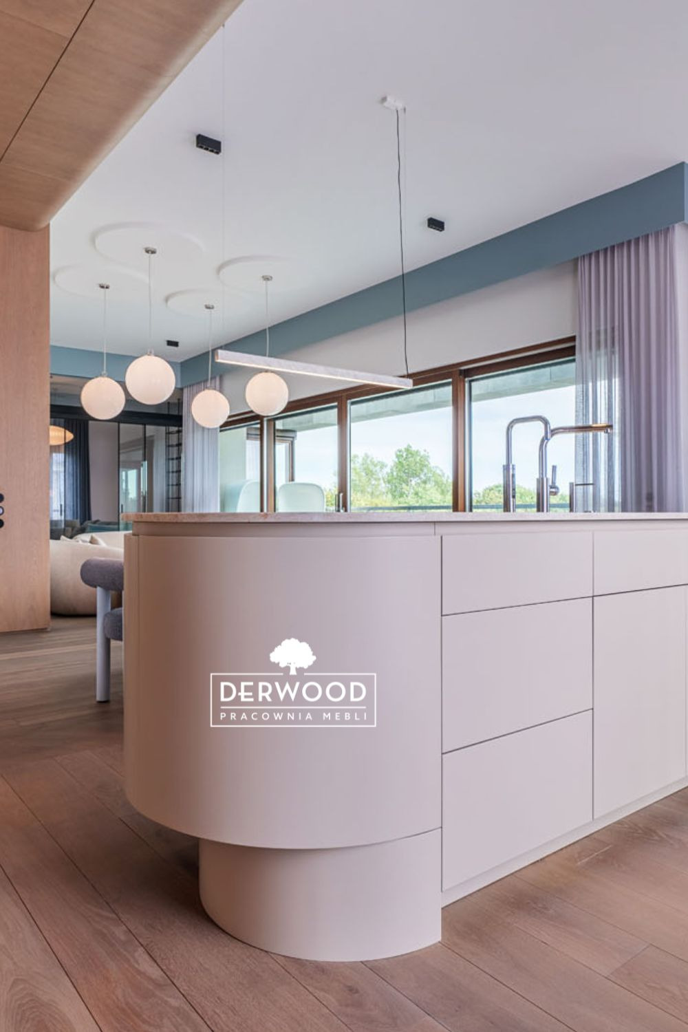 Wyspa kuchenna z zaokrąglonym frontem w odcieniach beżu, z widocznym logo firmy meblarskiej, w tle okno, wiszące lampy i fragment salonu.