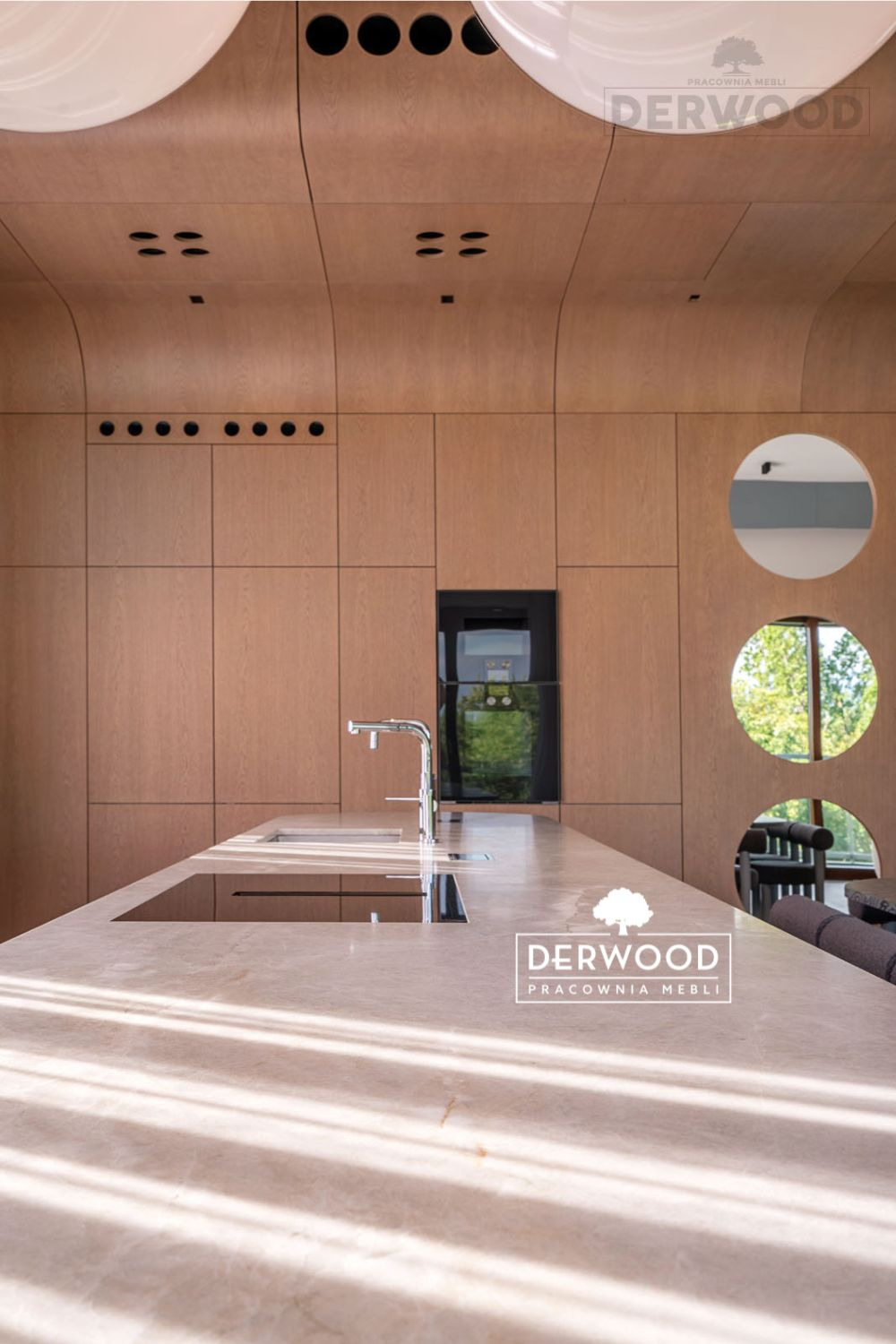 Nowoczesna kuchnia z drewnianymi szafkami o jasnym odcieniu, okrągłymi oknami i wyspą z marmurowym blatem, oświetlona promieniami słońca.