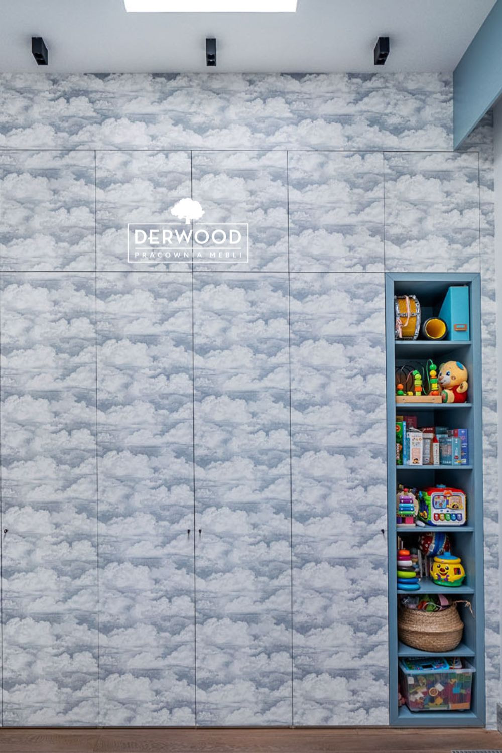 Szafa w pokoju dziecięcym z frontami pokrytymi tapetą w chmury, obok regał z niebieskimi półkami wypełniony zabawkami. Na szafie widoczne logo firmy meblarskiej Derwood.