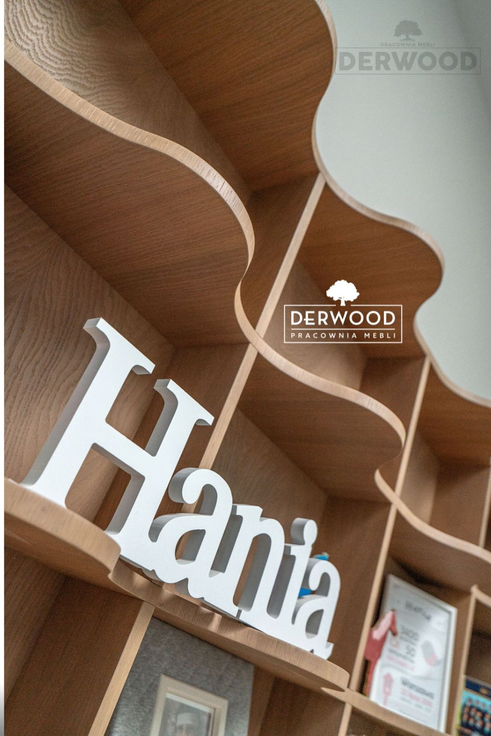 Półki regału z litego drewna o falistych kształtach, na jednej z półek biały napis 3D 'Hania' oraz ramki ze zdjęciami, widoczne logo firmy meblarskiej w rogu.