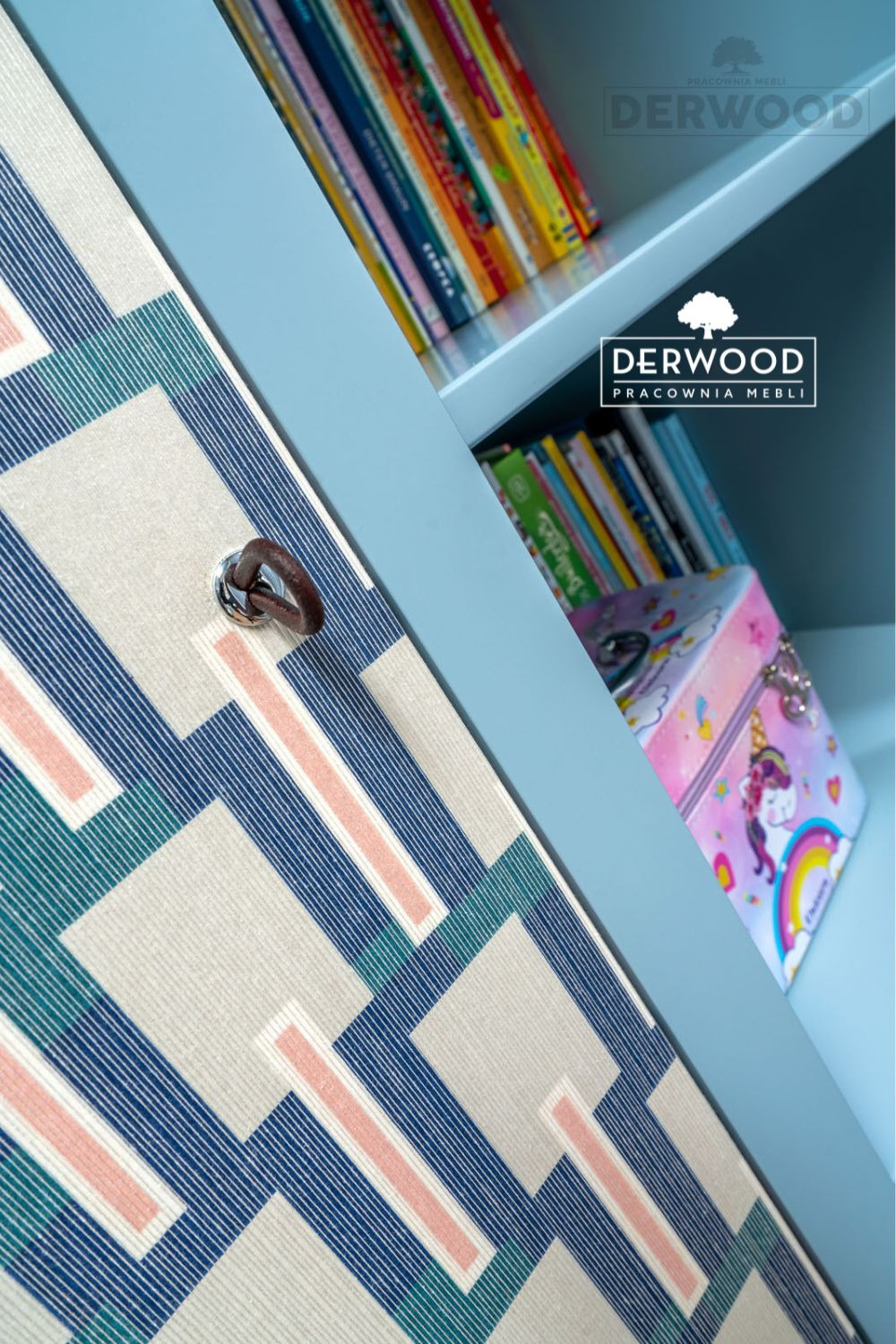 Niebieski regał z geometrycznym wzorem na drzwiach, skórzany uchwyt i półki wypełnione książkami oraz pudełkiem z jednorożcem.