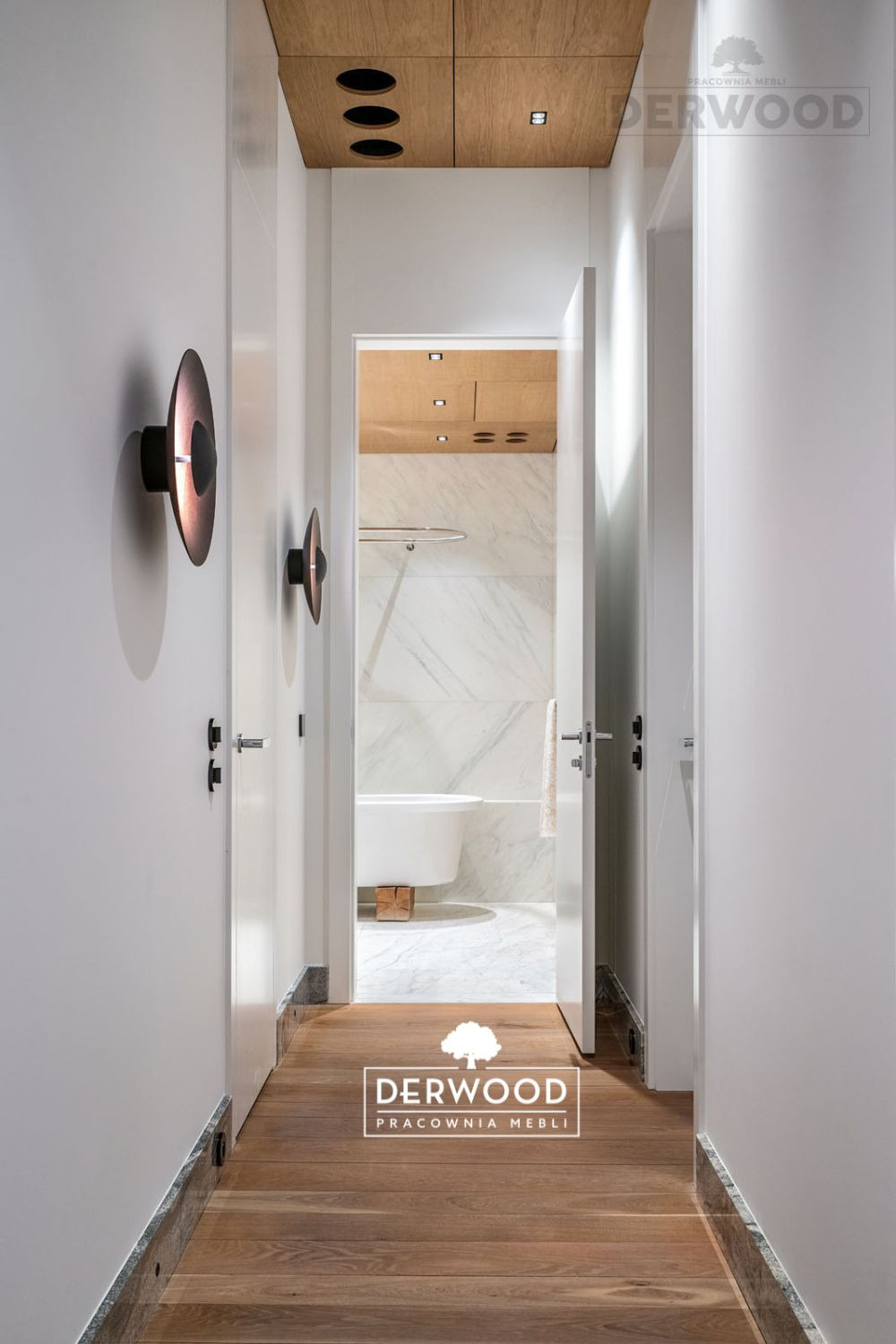 Wnętrze korytarza z drewnianą podłogą i widokiem na łazienkę z białą wanną na drewnianym podeście. Na ścianie okrągłe lampy. Zabudowa sufitowa z drewna z okrągłymi otworami wentylacyjnymi.