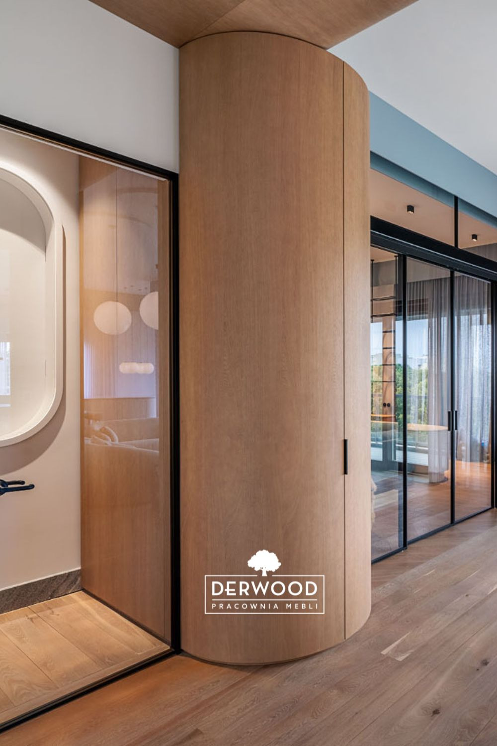 Nowoczesne wnętrze z zaokrąglonym, drewnianym słupem/szafą w zabudowie, szklane drzwi przesuwne z czarnymi ramami, parkiet w ciepłym odcieniu i logo firmy meblarskiej w dolnej części.