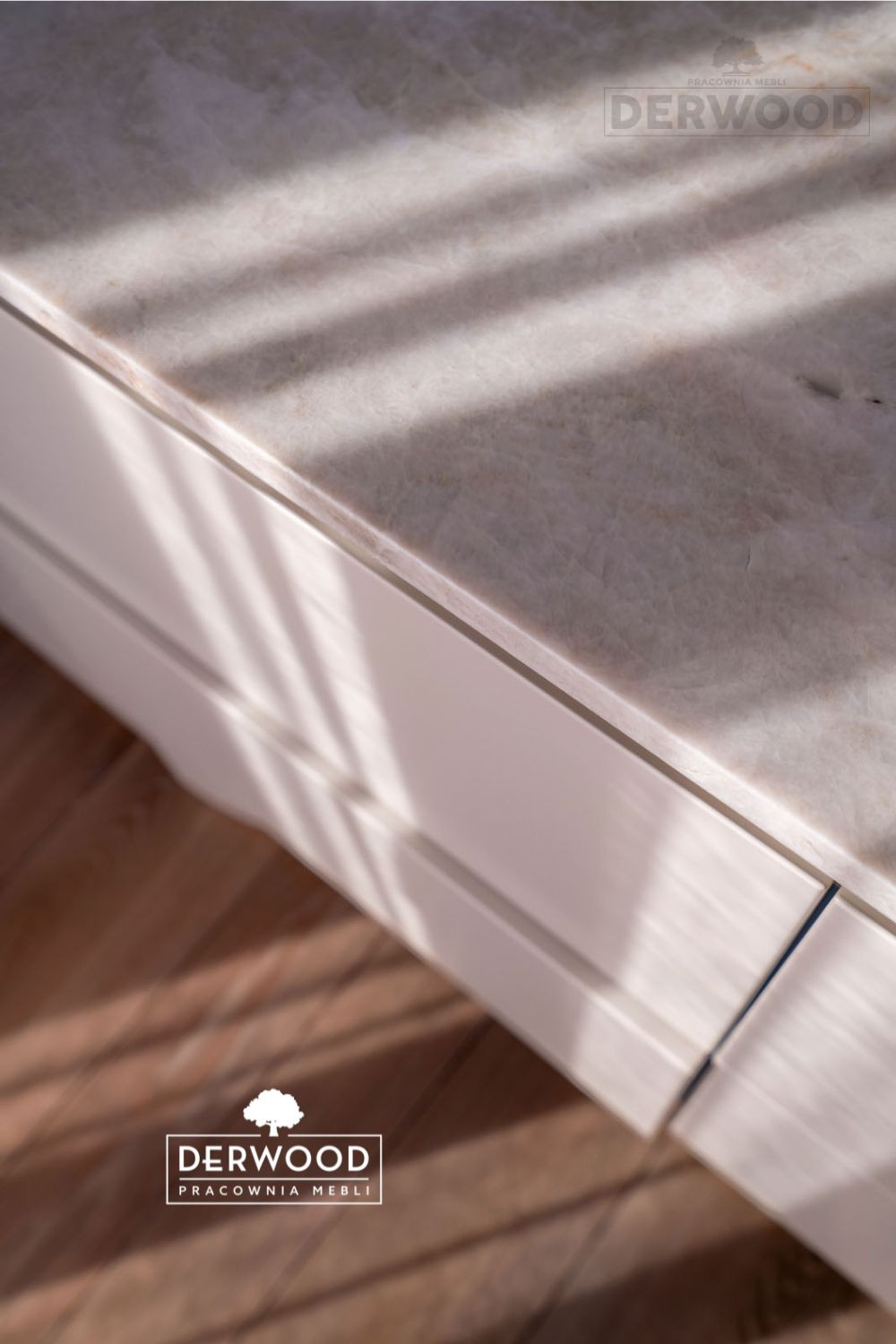 Fragment białej zabudowy kuchennej z marmurowym blatem, oświetlonej promieniami słonecznymi, z widocznym logo firmy meblarskiej.