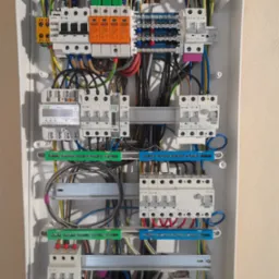 Rozdzielnica elektryczna z zainstalowanymi bezpiecznikami, licznikiem i przewodami w kolorach: niebieskim, żółtym, zielonym, czarnym i czerwonym w białej obudowie.