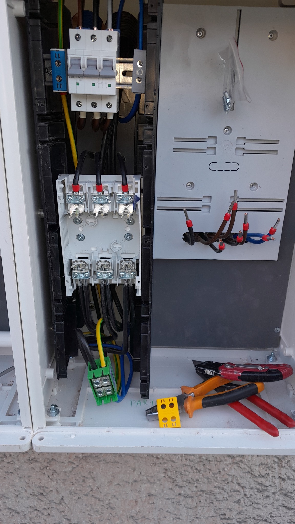 Otwarta skrzynka elektryczna z widocznymi przewodami, bezpiecznikami i narzędziami elektrycznymi u dołu. Widoczne złącza kablowe i zaciski.