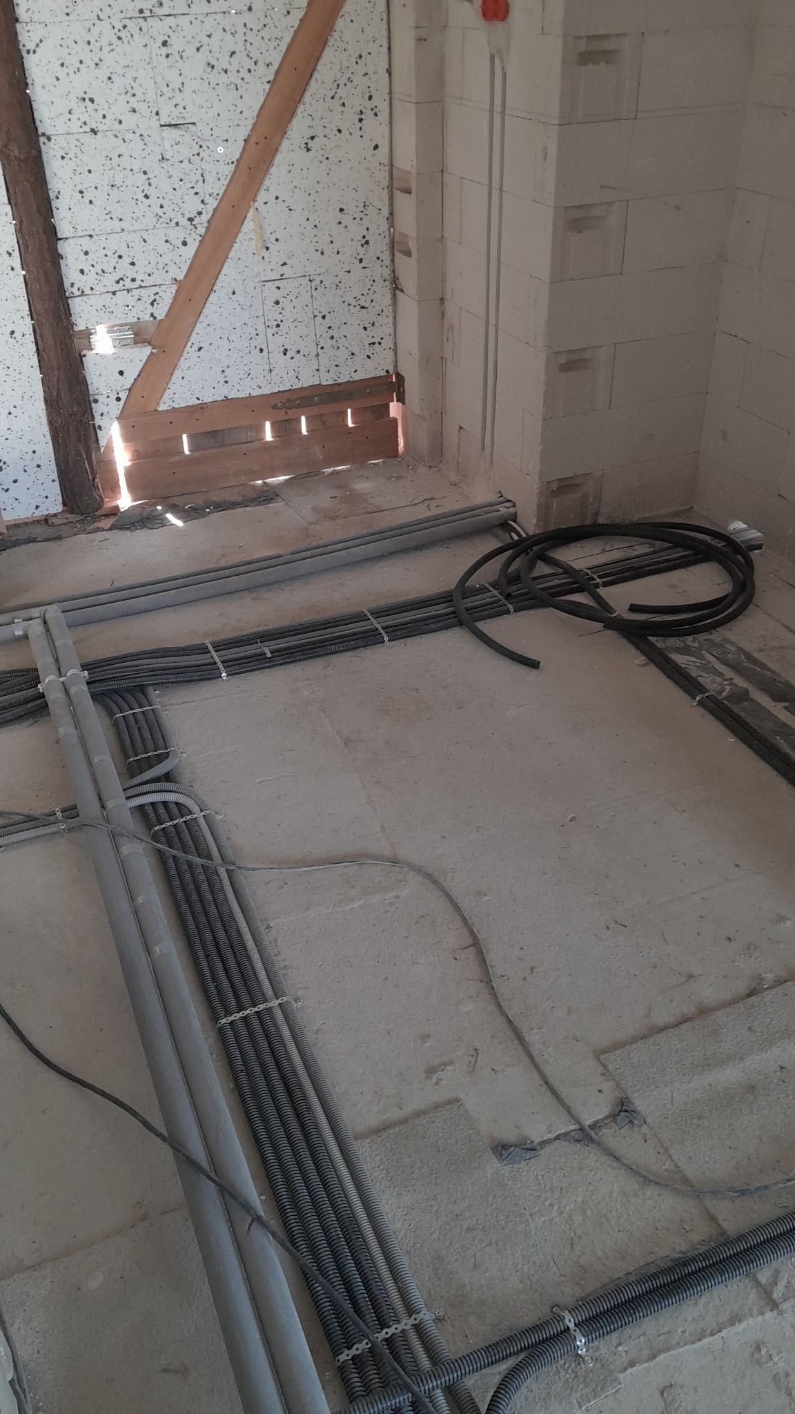 Szare rury i peszle elektryczne ułożone na surowym podłożu wewnątrz budynku w trakcie instalacji. Widoczne ściany z pustaków i fragmenty drewnianej konstrukcji.