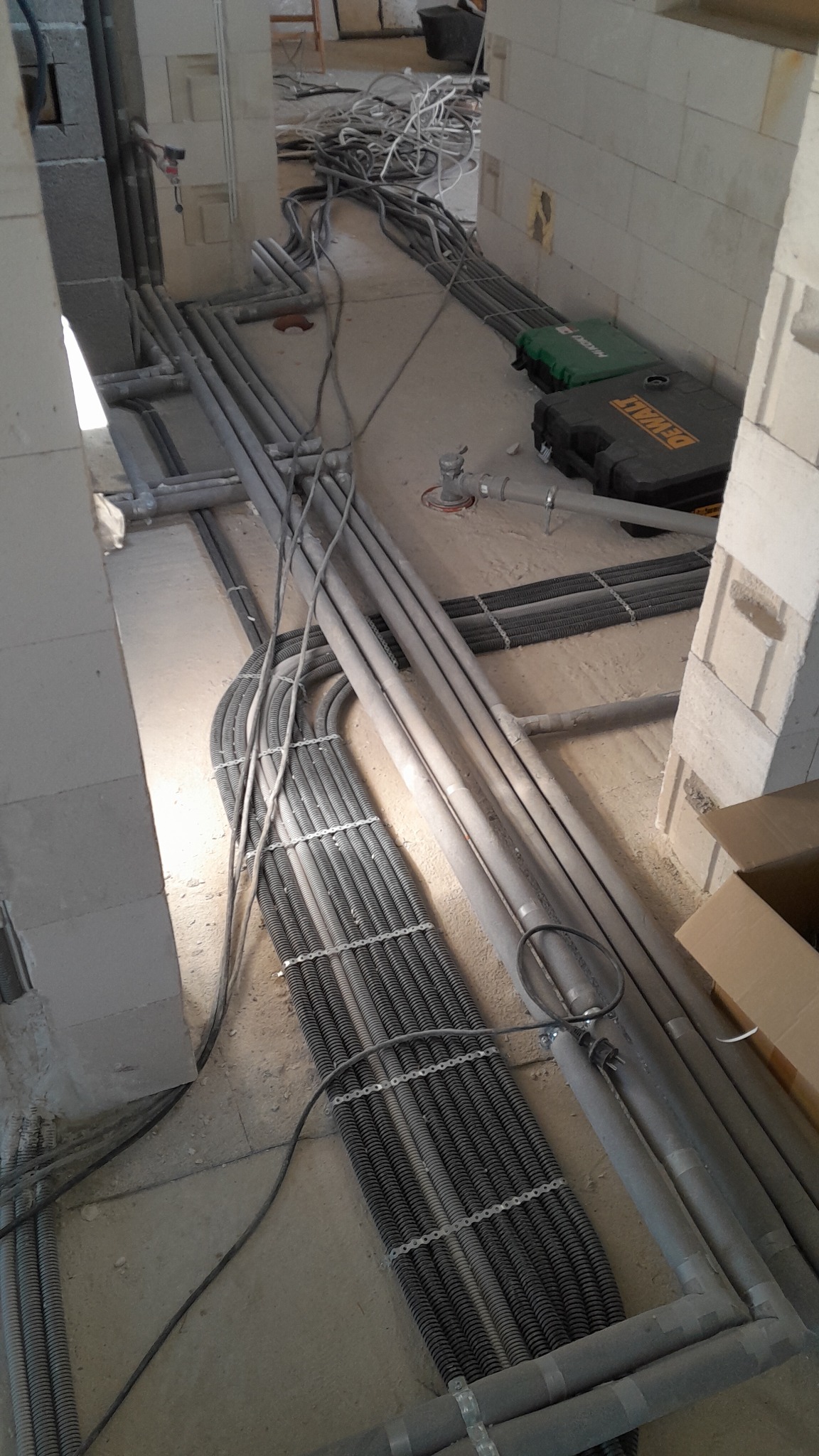 Szara instalacja elektryczna w rurach PCV i peszlach, ułożona w surowym wnętrzu budynku w trakcie prac instalacyjnych. Widoczne narzędzia w tle.