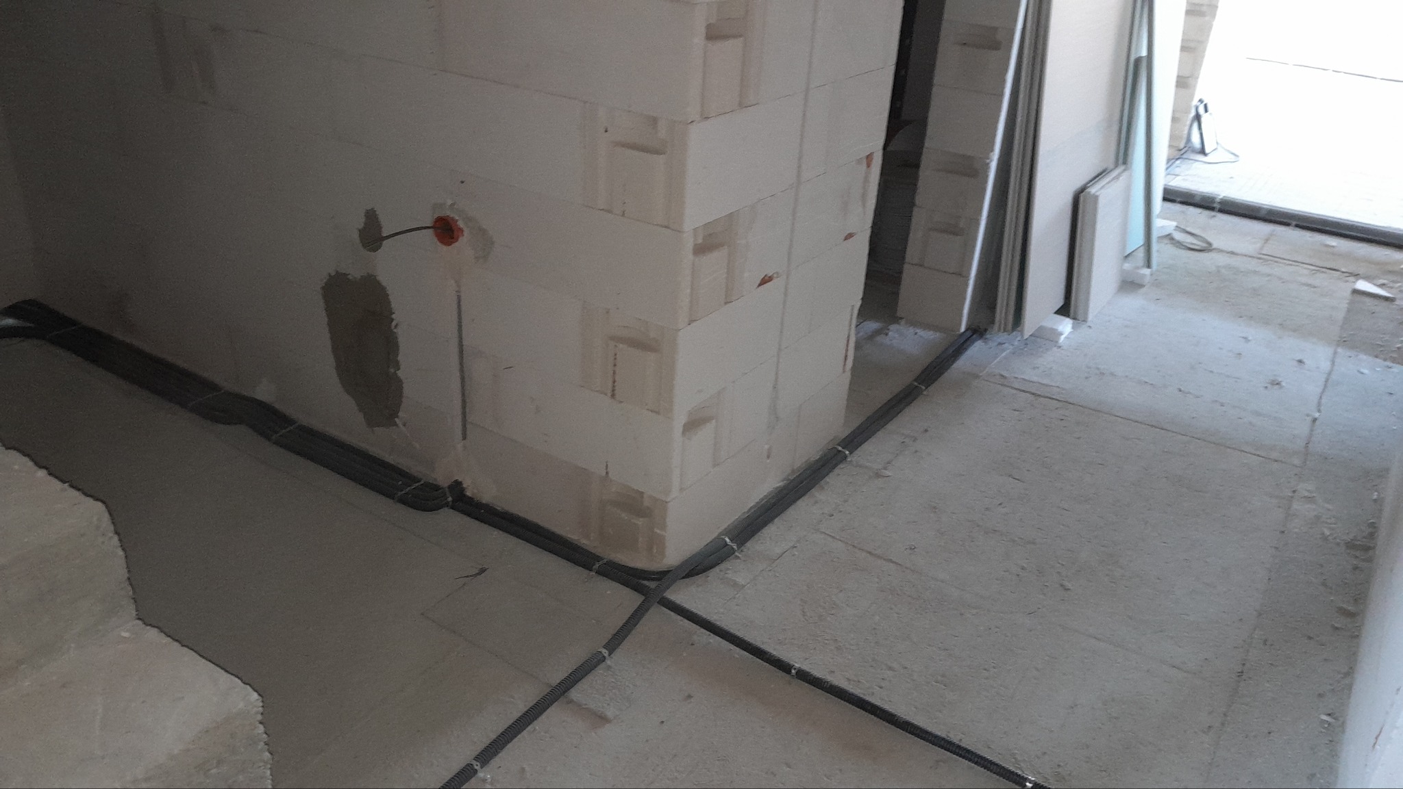 Instalacja przewodów w peszlach przy ścianie z betonu komórkowego w surowym wnętrzu. Widoczna puszka elektryczna i fragment podłogi.