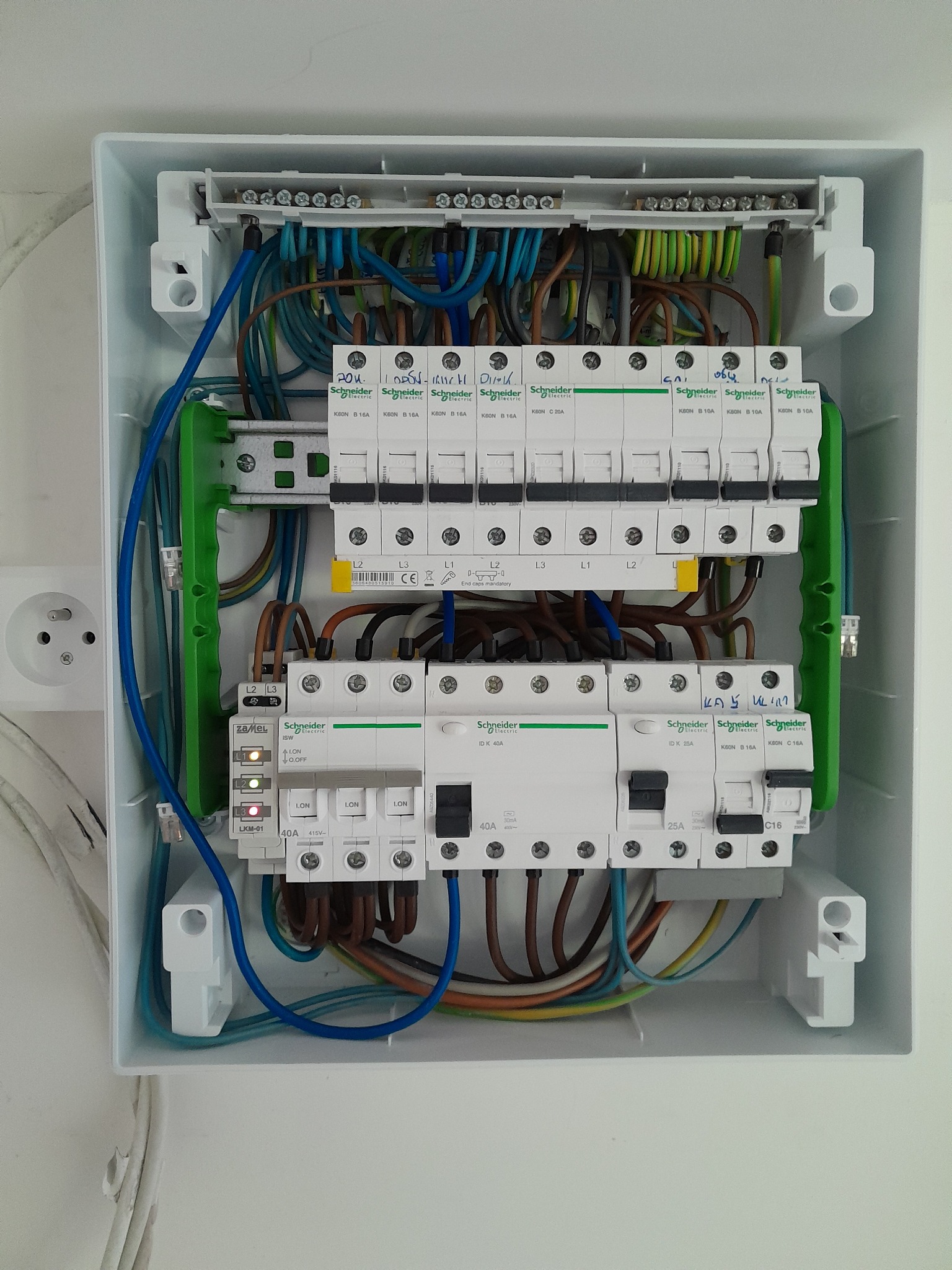 Rozdzielnica elektryczna w białej obudowie, z widocznymi bezpiecznikami, przewodami różnej barwy i elementami montażowymi. Widoczne oznaczenia Schneider Electric.