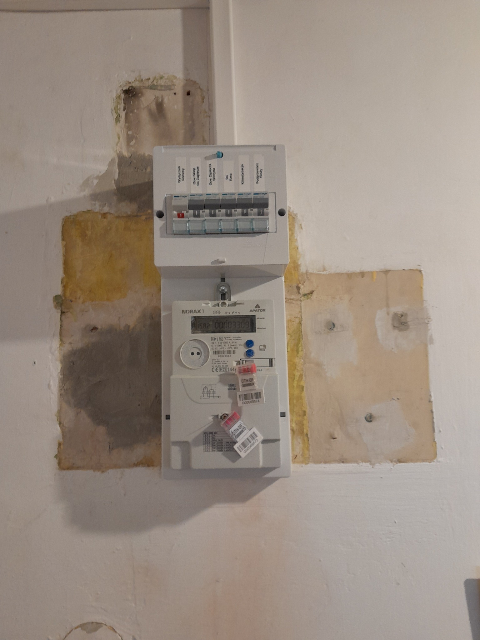 Zamontowany licznik elektryczny NORAX1 na ścianie z widocznymi śladami po instalacji, otoczony łatami gipsowymi. Widoczne bezpieczniki i wyświetlacz licznika.