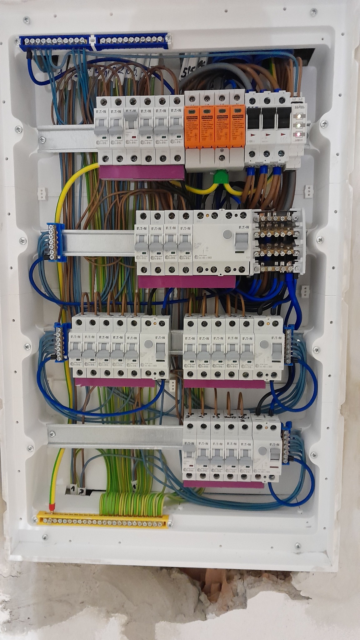Rozdzielnia elektryczna w białej obudowie z widocznymi bezpiecznikami, przewodami i listwami zaciskowymi. Instalacja w trakcie montażu, widoczne kable i elementy zabezpieczeń.
