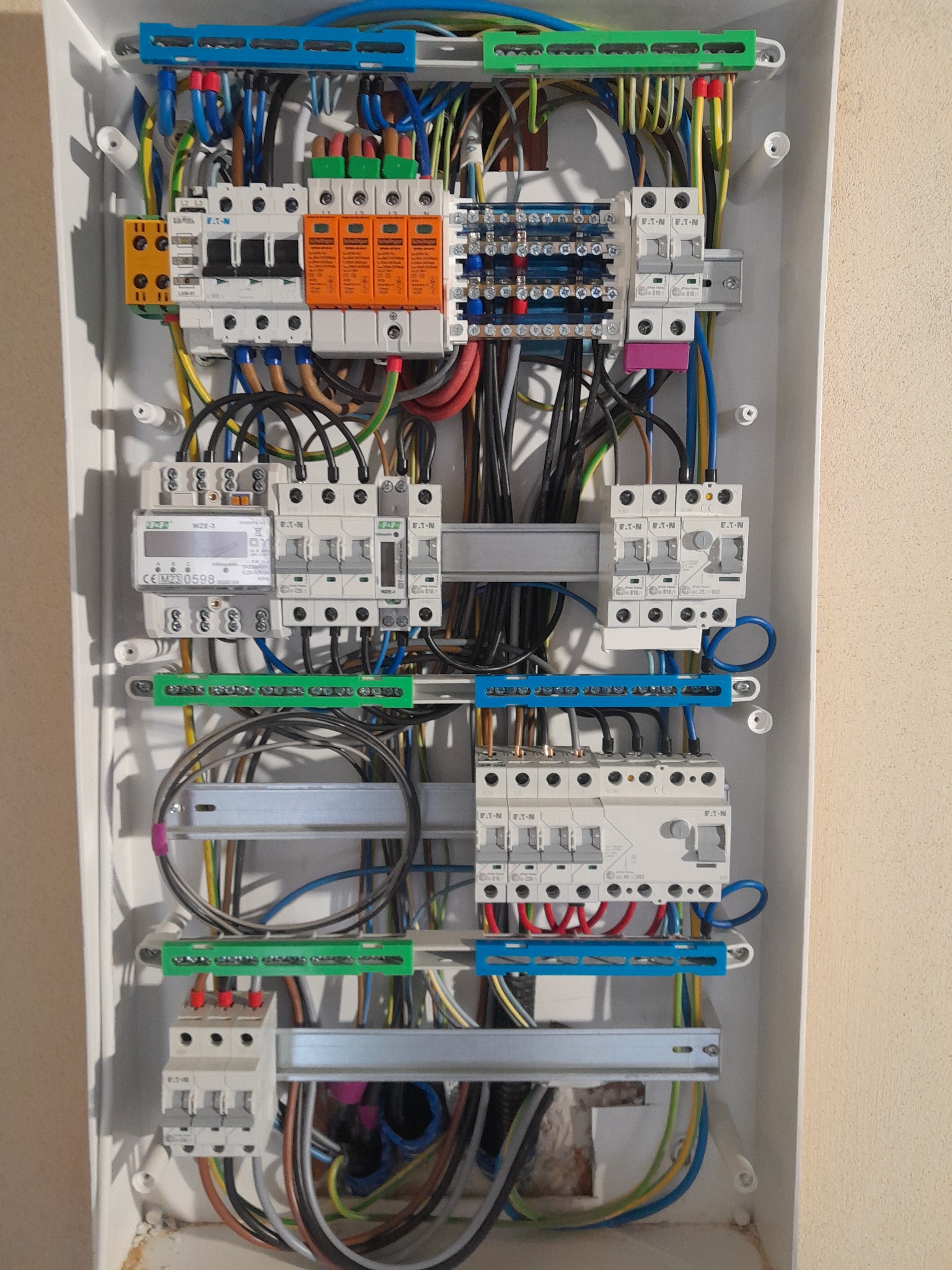 Rozdzielnica elektryczna z zainstalowanymi bezpiecznikami, licznikiem i przewodami w kolorach: niebieskim, żółtym, zielonym, czarnym i czerwonym w białej obudowie.