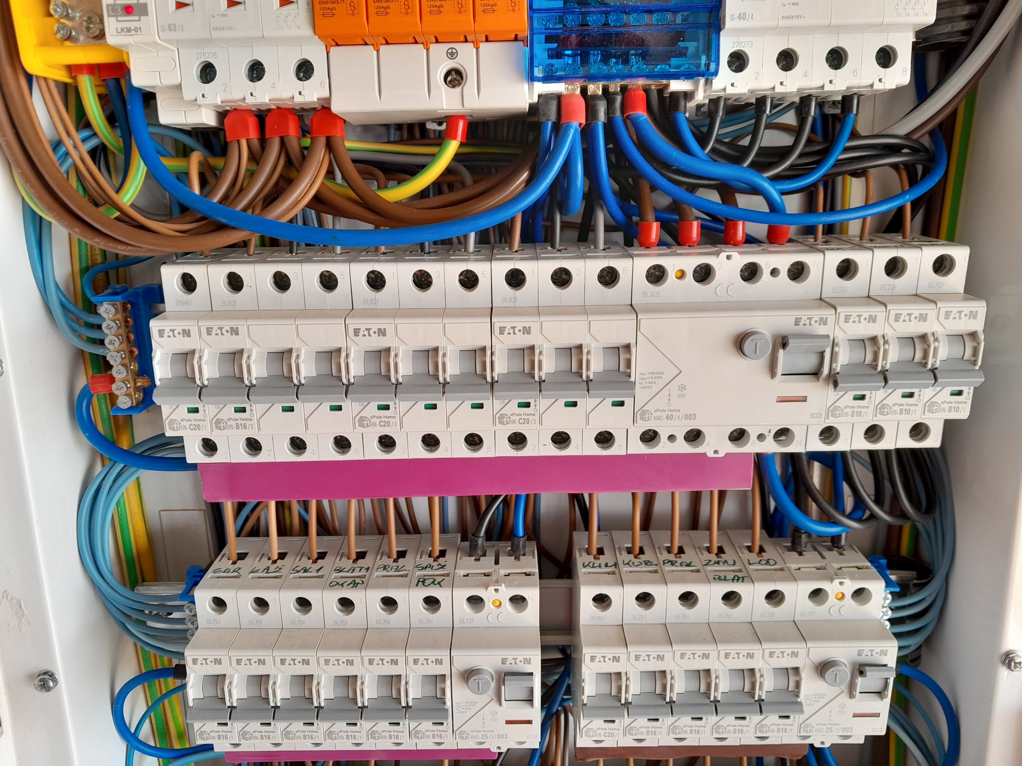 Rozdzielnica elektryczna z bezpiecznikami EATON, okablowaniem w kolorach niebieskim, brązowym i żółto-zielonym. Widoczne opisy obwodów na listwach.
