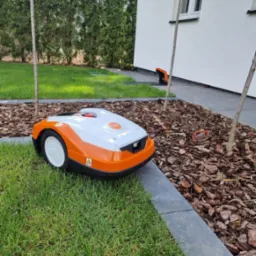 Pomarańczowo-biały robot koszący Stihl na trawniku obok rabaty z korą, w tle budynek z białymi ścianami i zielone tuje.