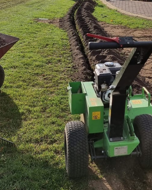 Wąski wykop liniowy w trawniku wykonany specjalistyczną maszyną, obok taczka z ziemią.