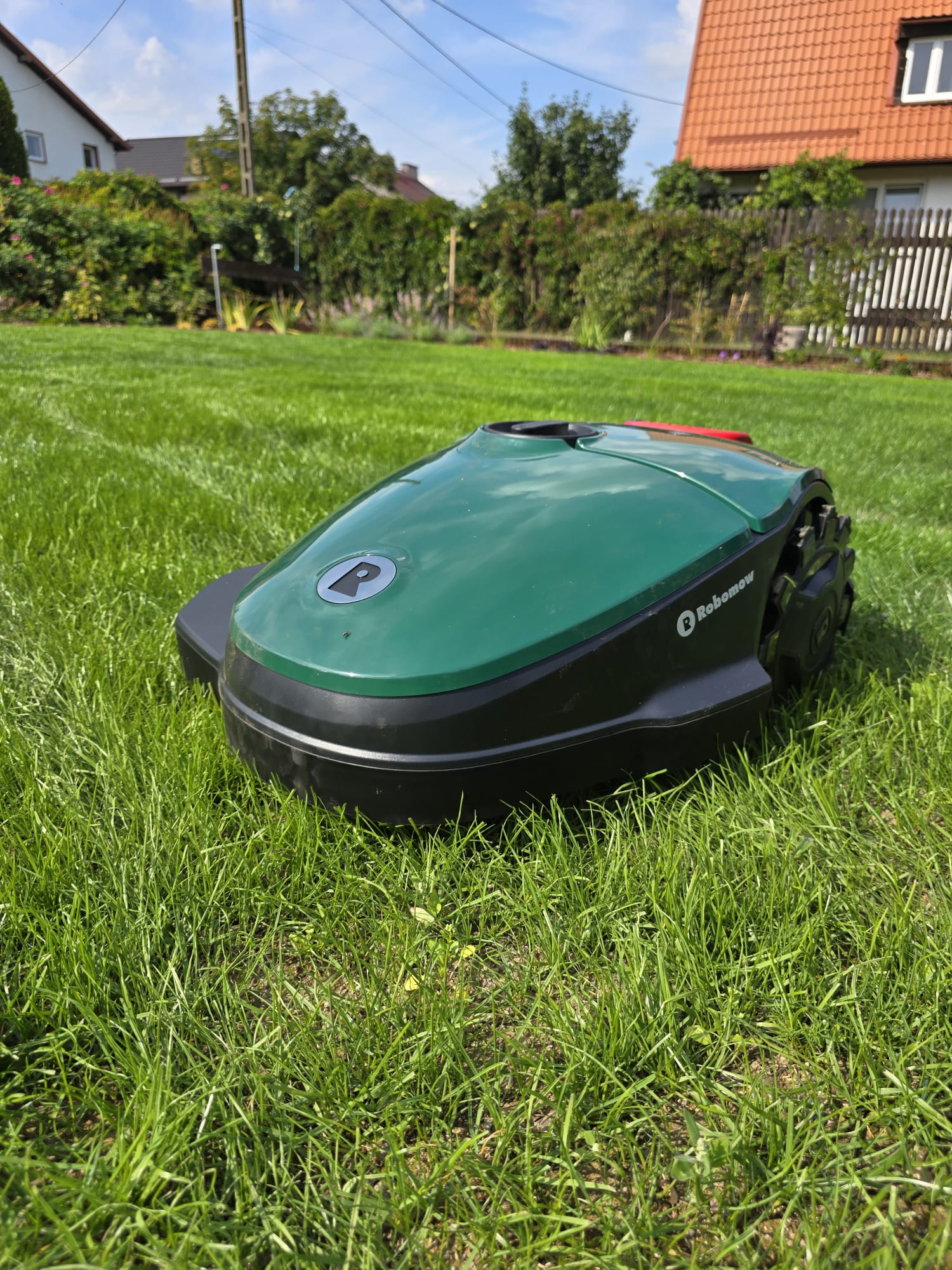 Zielony robot koszący Robomow na idealnie przystrzyżonym trawniku przed domem z czerwonym dachem i białym ogrodzeniem w słoneczny dzień.