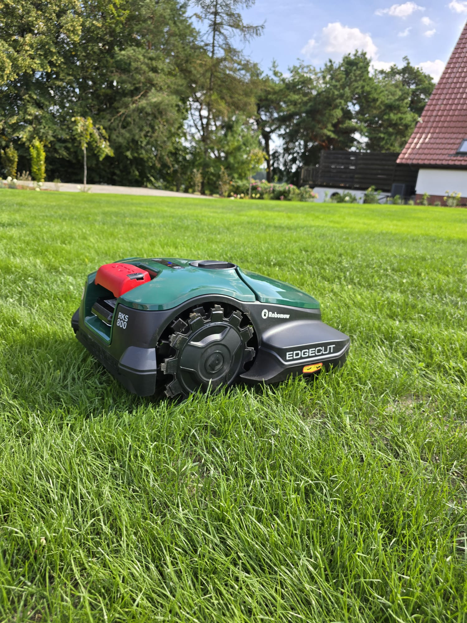 Robot koszący Robomow RKS 800 na zadbanym trawniku przed domem z czerwonym dachem, widok z niskiej perspektywy.