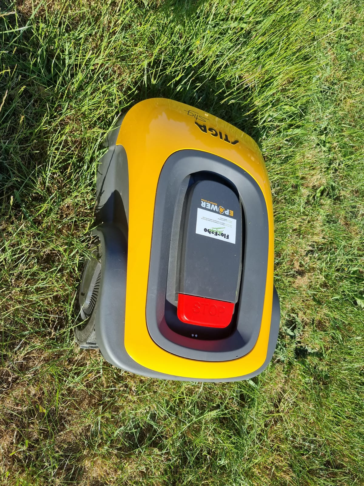 Żółto-szary robot koszący Stiga na trawniku, widoczny z góry, z bliska. Czerwony przycisk STOP i etykieta z nazwą producenta Florfabo.