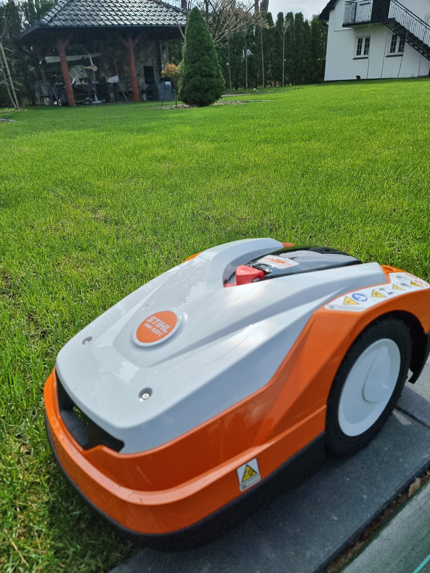 Automatyczna kosiarka Stihl na tle wypielęgnowanego trawnika przed domem z altaną w ogrodzie.