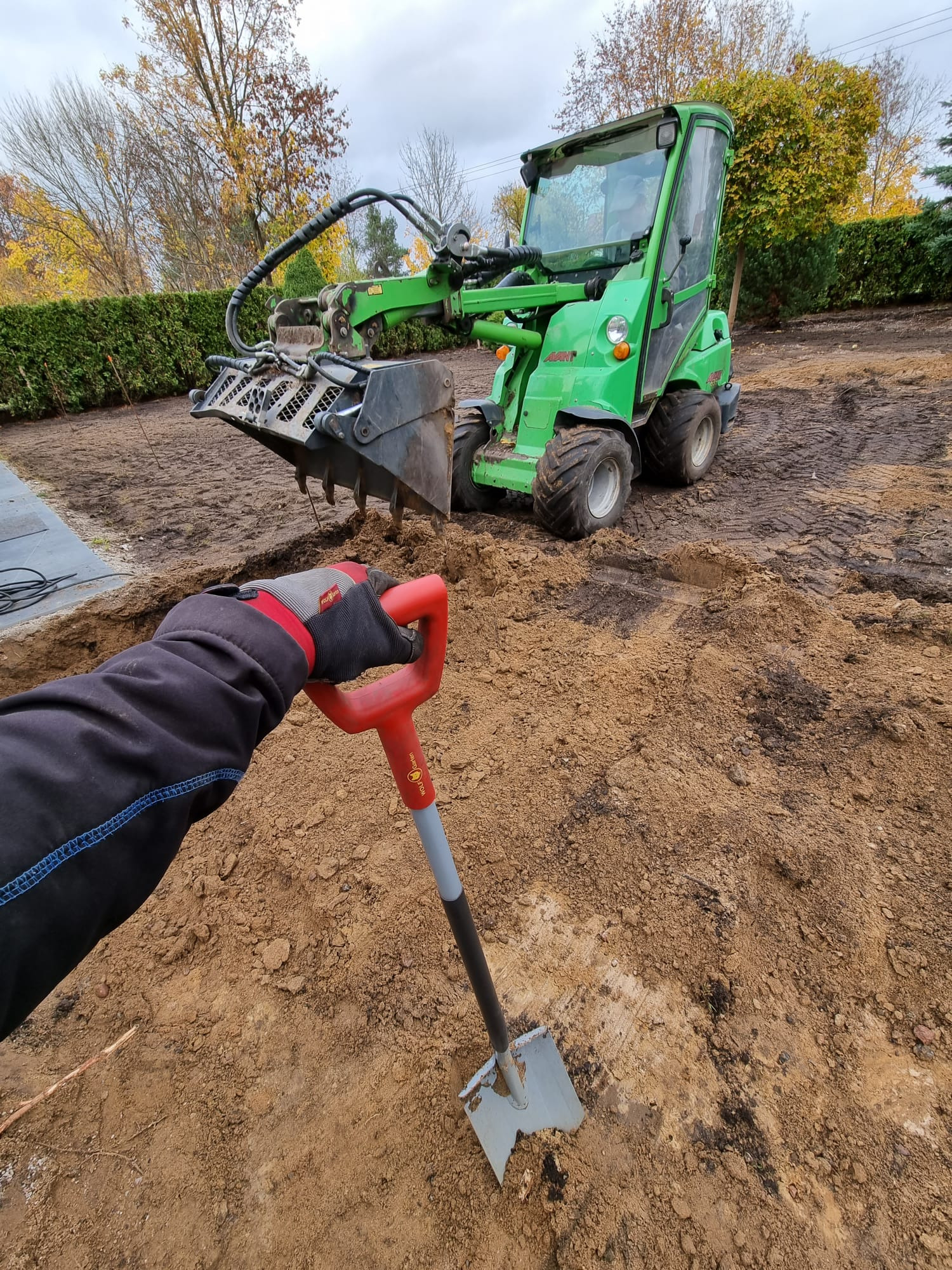 Ręka w roboczej rękawicy trzyma szpadel na tle wykopu, z zieloną mini-ładowarką Avant w tle, przygotowanie terenu pod ogród.