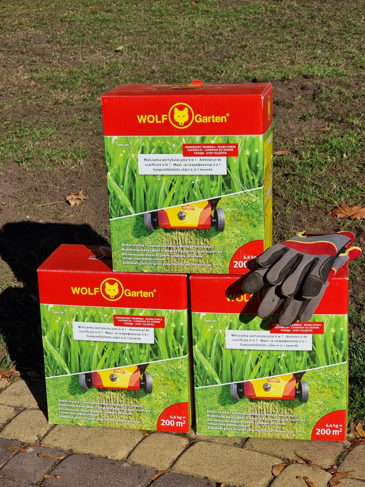 Trzy opakowania mieszanki wertykulacyjnej 4 w 1 firmy Wolf Garten na tle kostki brukowej i trawnika, obok leżą robocze rękawice, zdjęcie wykonane w Mrągowie.