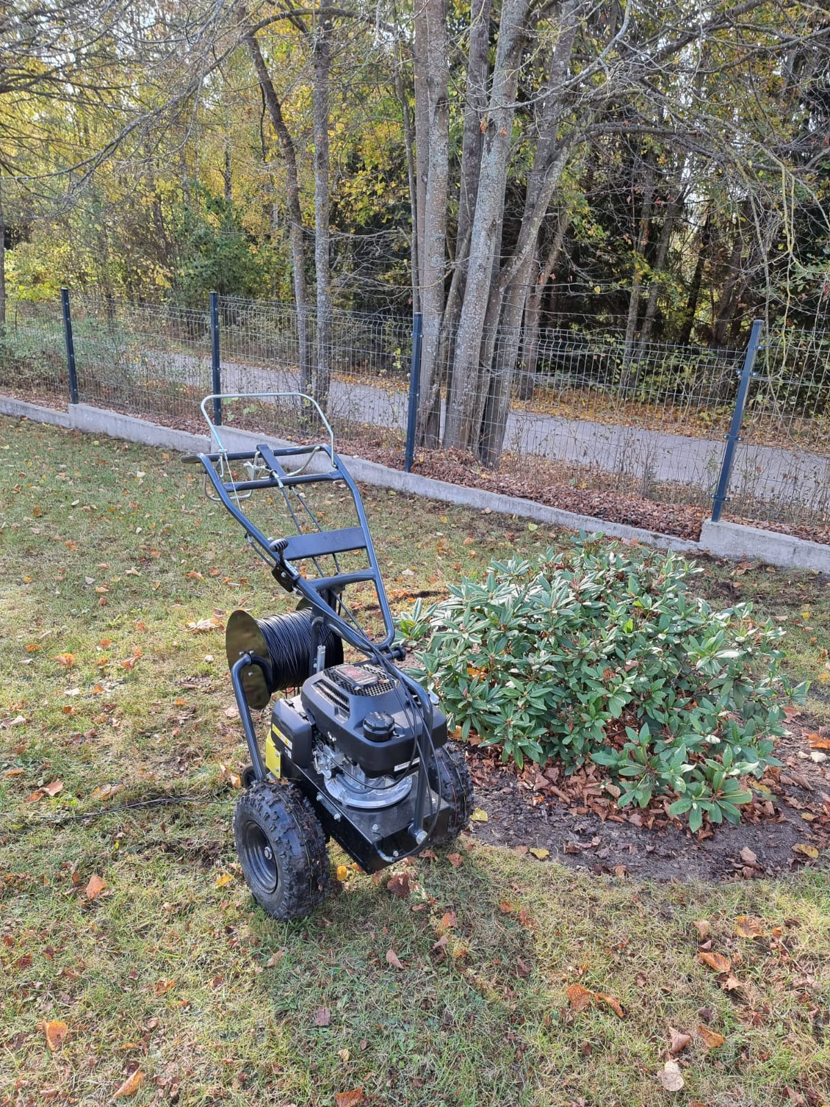 Profesjonalna maszyna do wykopów pod system nawadniania z nawiniętym przewodem, stojąca na trawniku w ogrodzie, z widocznym krzewem i ogrodzeniem w tle.