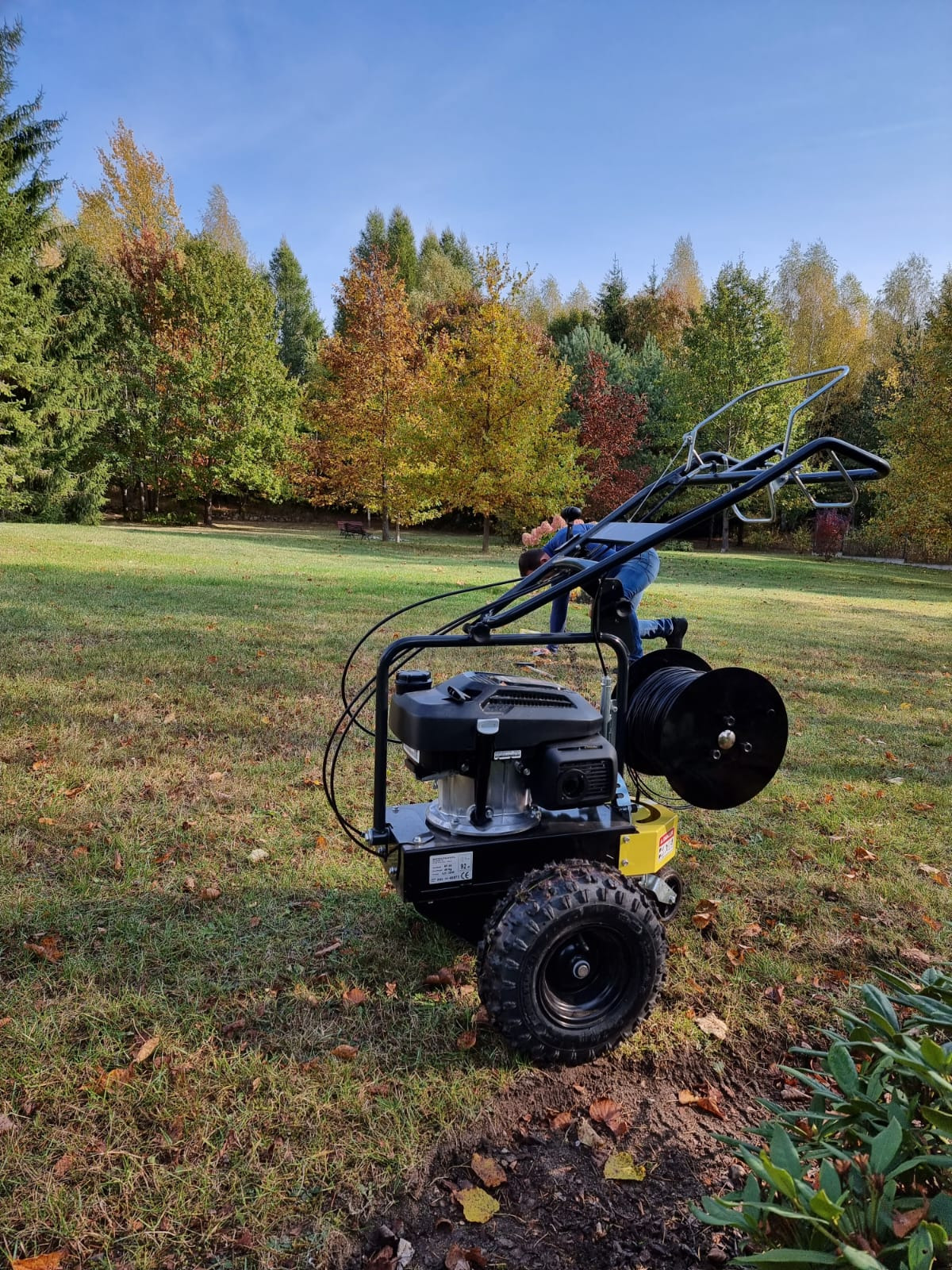 Maszyna do wykopów pod instalacje nawadniające na trawniku, widoczna osoba obsługująca urządzenie, jesienne drzewa w tle.