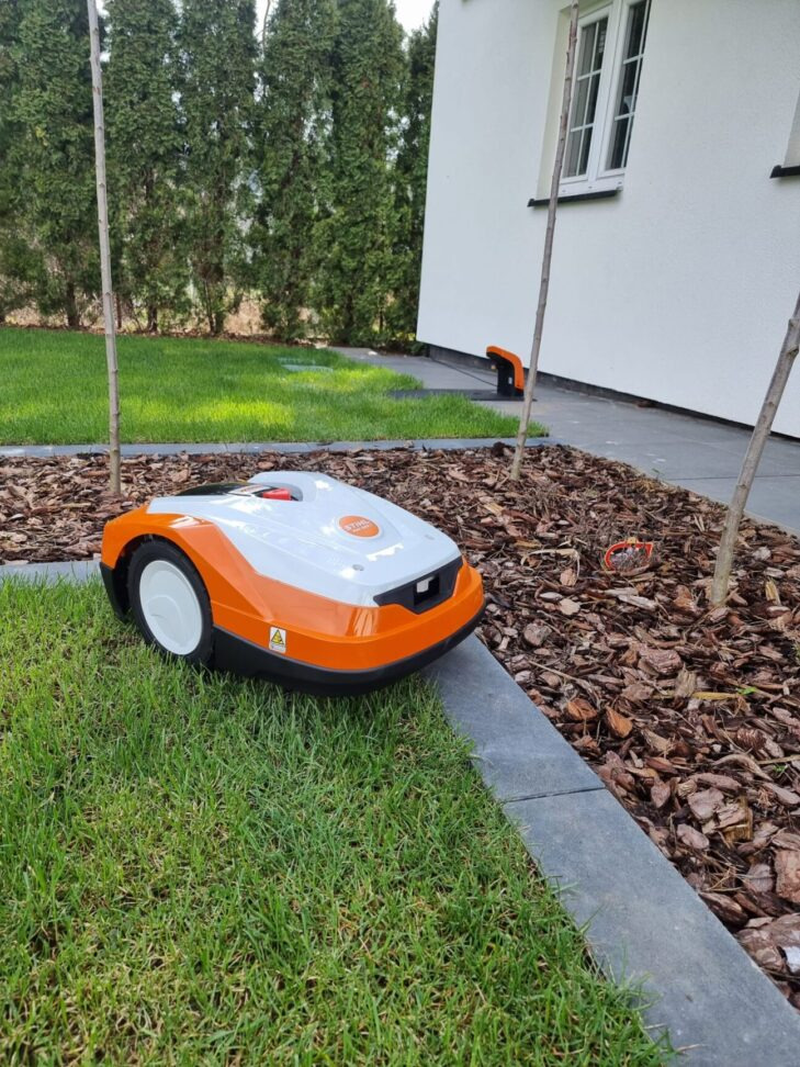 Pomarańczowo-biały robot koszący Stihl na trawniku obok rabaty z korą, w tle budynek z białymi ścianami i zielone tuje.