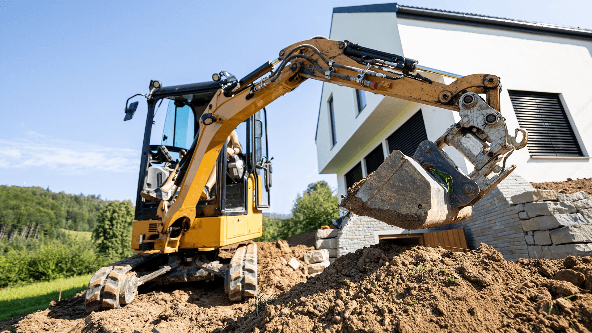 Żółta mini koparka w trakcie prac ziemnych, nabiera ziemię z wykopu, w tle nowoczesny dom z szarymi roletami.