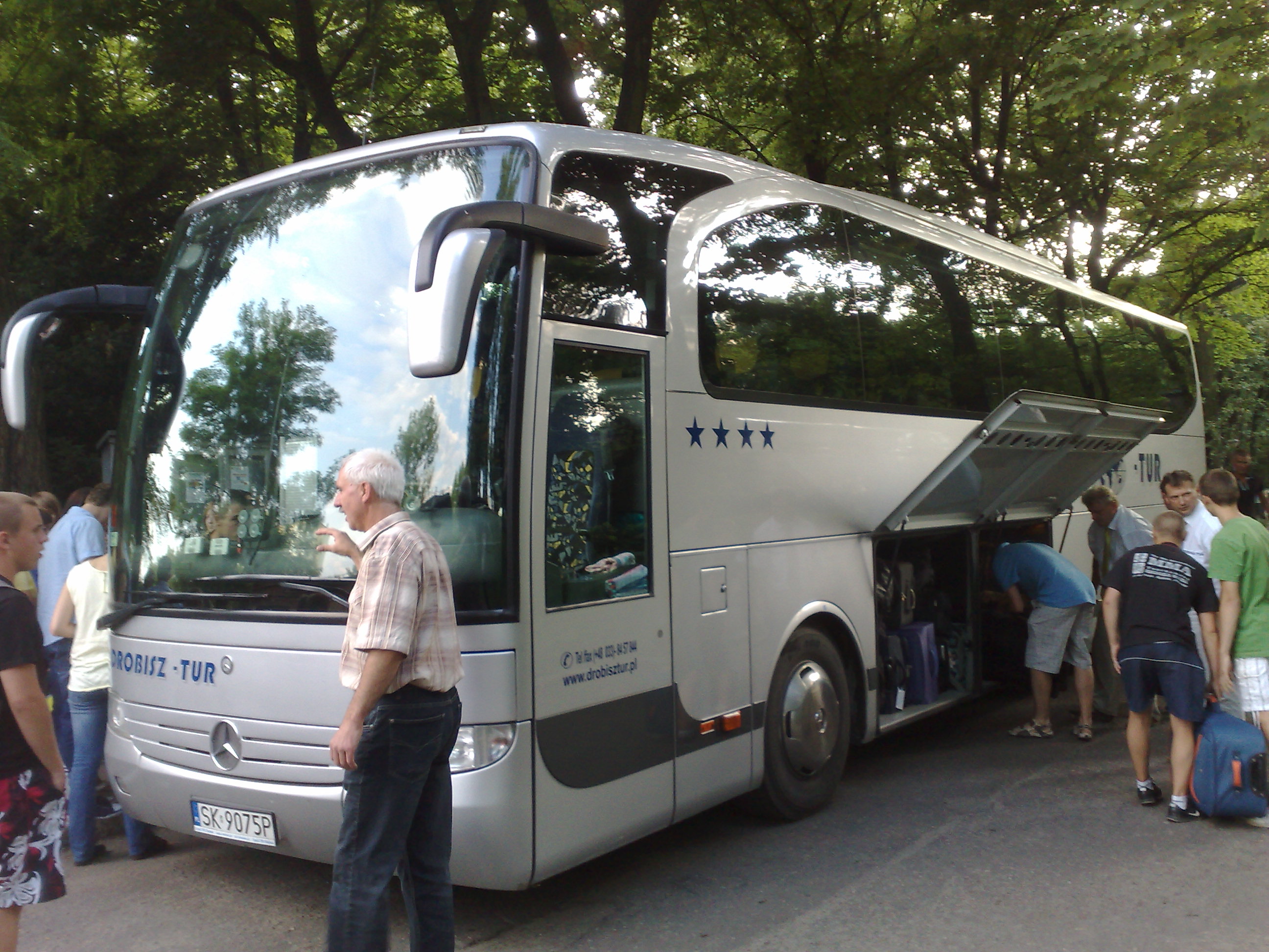 Srebrny autokar turystyczny marki Mercedes z otwartym lufcikiem bagażowym, pasażerowie wkładają bagaże, widoczny kierowca w koszuli w kratę, tablica rejestracyjna SK 9075P i logo firmy Robisz-Tur.