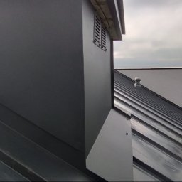 Robert Jurczak - Szary dach z blachy trapezowej z kominem i wentylacją, na tle krajobrazu. Widoczne detale obróbek blacharskich i łączeń.