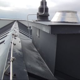 Robert Jurczak - Metalowy dach z kominem i systemem odgromowym na tle krajobrazu. Widoczne detale montażu i obróbki blacharskie.