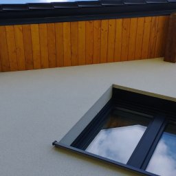 Robert Jurczak - Fragment elewacji domu z oknem i drewnianą podbitką dachową z widocznymi belkami. Czarna rynna wzdłuż krawędzi dachu. Jasny tynk strukturalny.