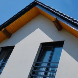 Nowoczesny dom z ciemną dachówką i drewnianą podbitką, widziany z dołu na tle błękitnego nieba. Wyraźne okna z czarnymi ramami i metalowymi barierkami.