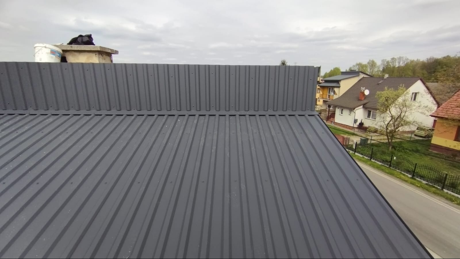 Szary dach z blachy trapezowej z widocznym kominem i elementami obróbki blacharskiej. W tle zabudowa jednorodzinna i pochmurne niebo.