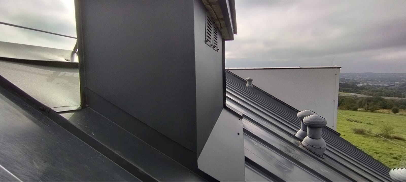 Szary dach z blachy trapezowej z kominem i wentylacją, na tle krajobrazu. Widoczne detale obróbek blacharskich i łączeń.