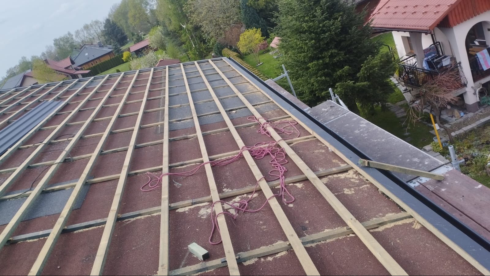 Dach w trakcie krycia: widoczne łaty, membrana i fragmenty blachy. Na dachu leży zwój liny. W tle domy i zieleń.