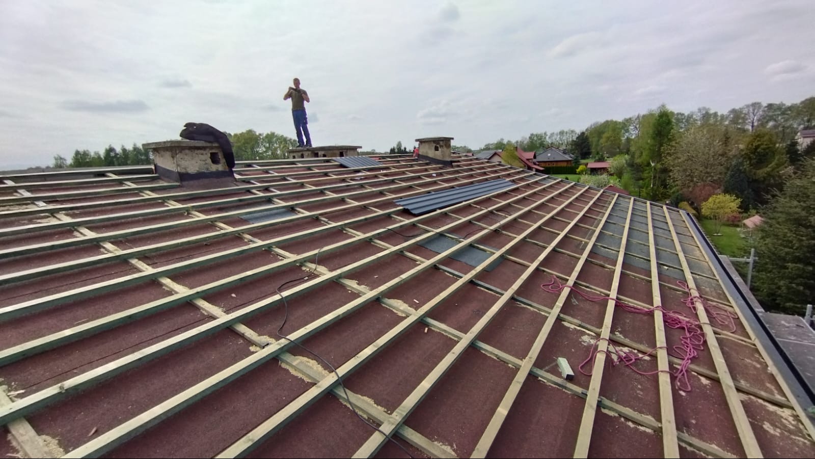 Konstrukcja dachu z łatami i kontrłatami, częściowo pokryta papą i blachodachówką. Na dachu stoi dekarz. Widok na okolicę.