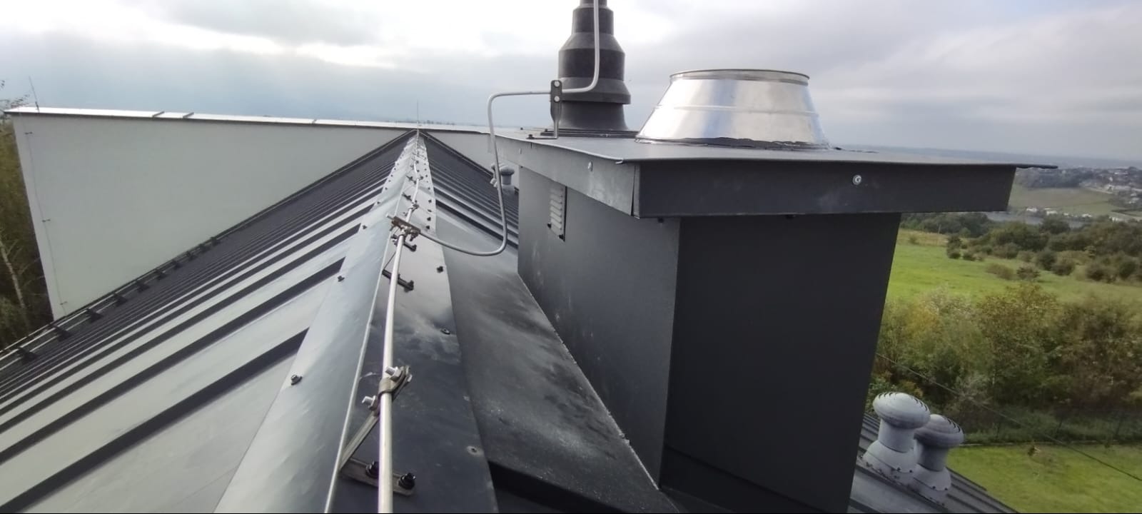 Metalowy dach z kominem i systemem odgromowym na tle krajobrazu. Widoczne detale montażu i obróbki blacharskie.